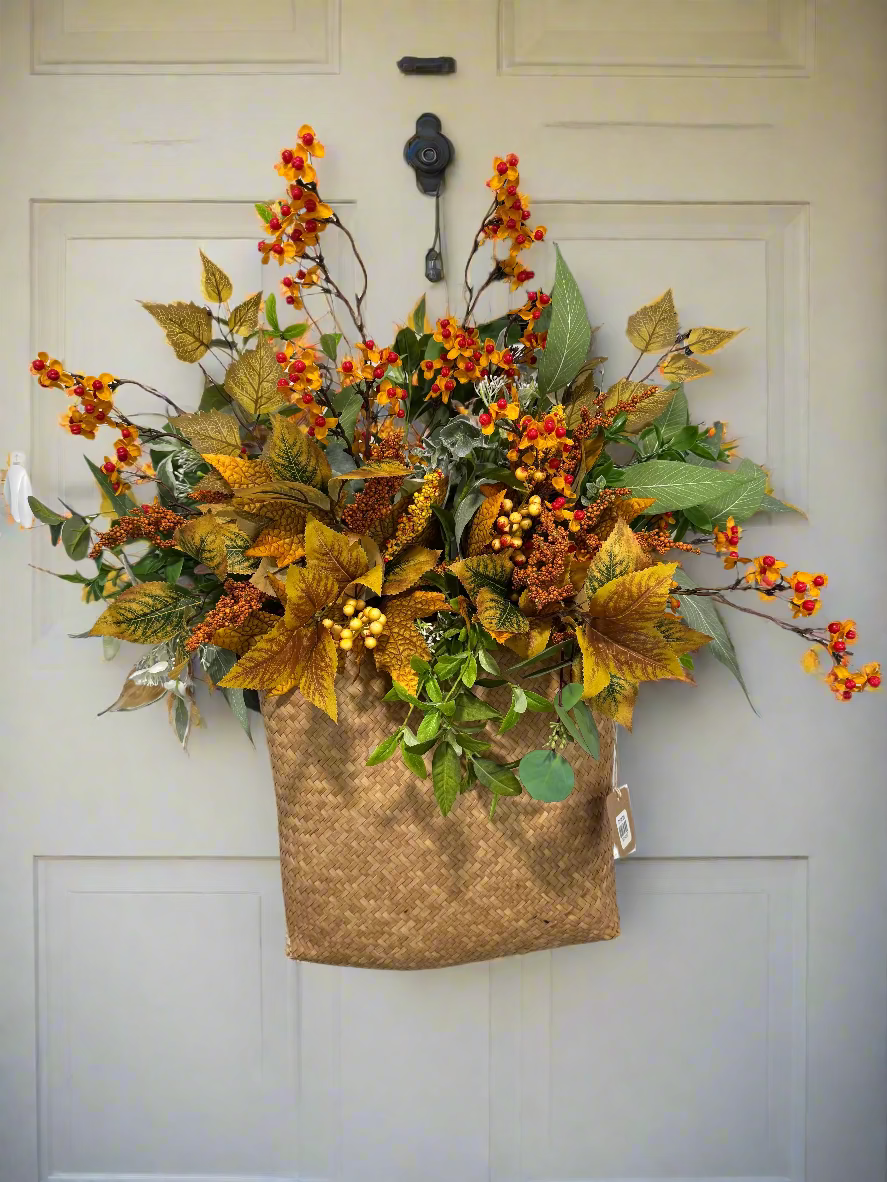 Artificial fall foliage arrangement in a woven basket on a white wall.