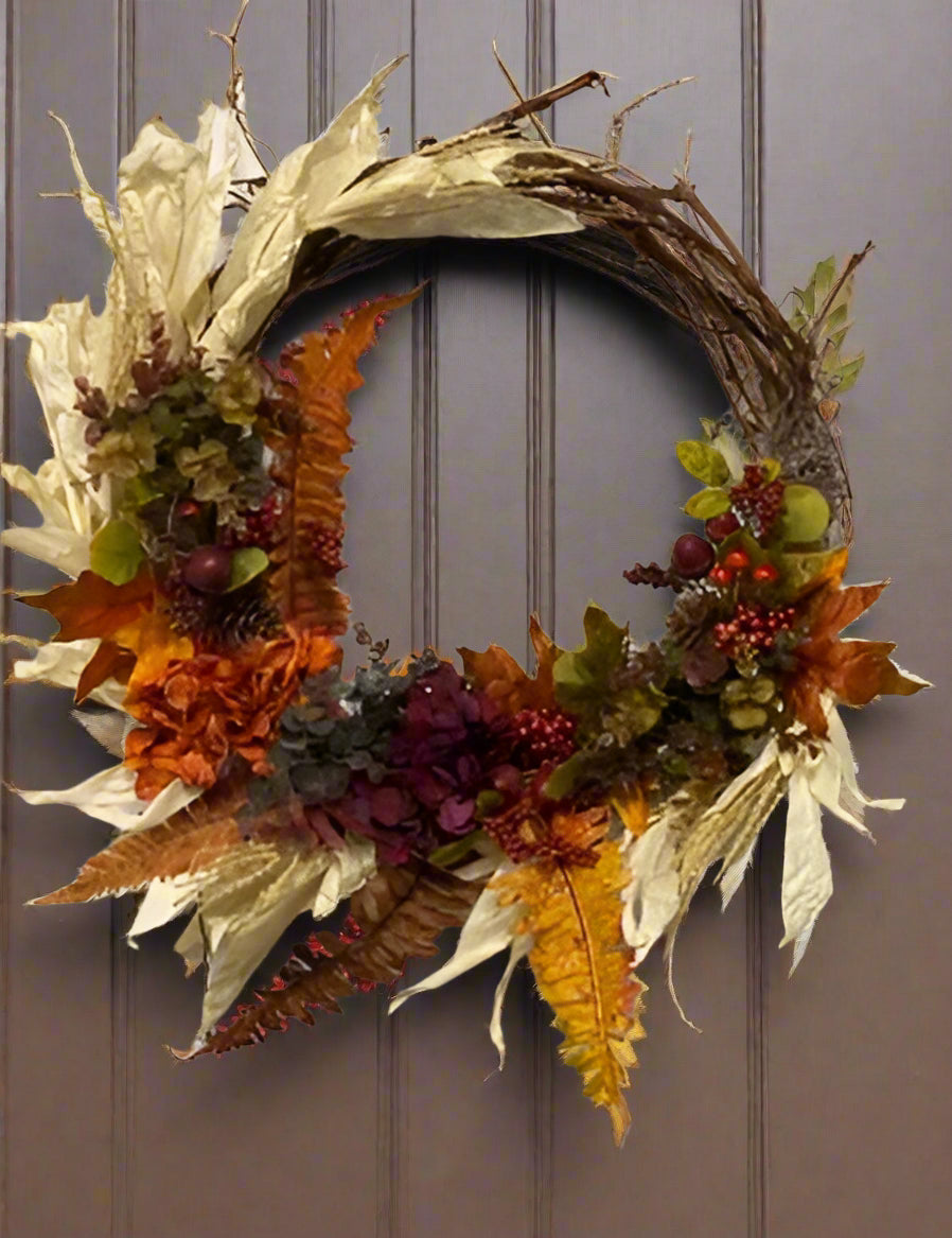 A fall wreath with a combination of orange, burgundy, and brown leaves, and decorative corn husks, displayed on a door.