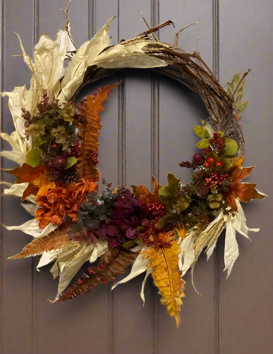 A fall wreath with a combination of orange, burgundy, and brown leaves, and decorative corn husks, displayed on a door.