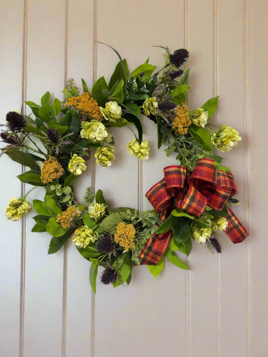 Decorative wreath with green leaves, yellow flowers, and a plaid ribbon on a white wall.