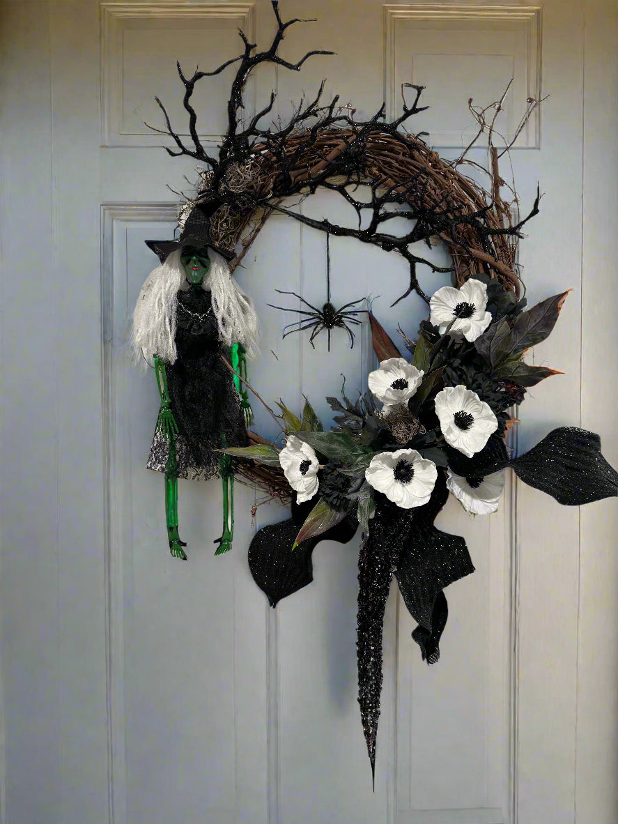 Halloween wreath with black and white flowers, branches, and a spider on a plain wall.