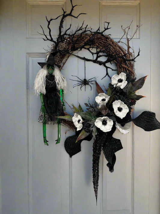Halloween wreath with black and white flowers, branches, and a spider on a plain wall.