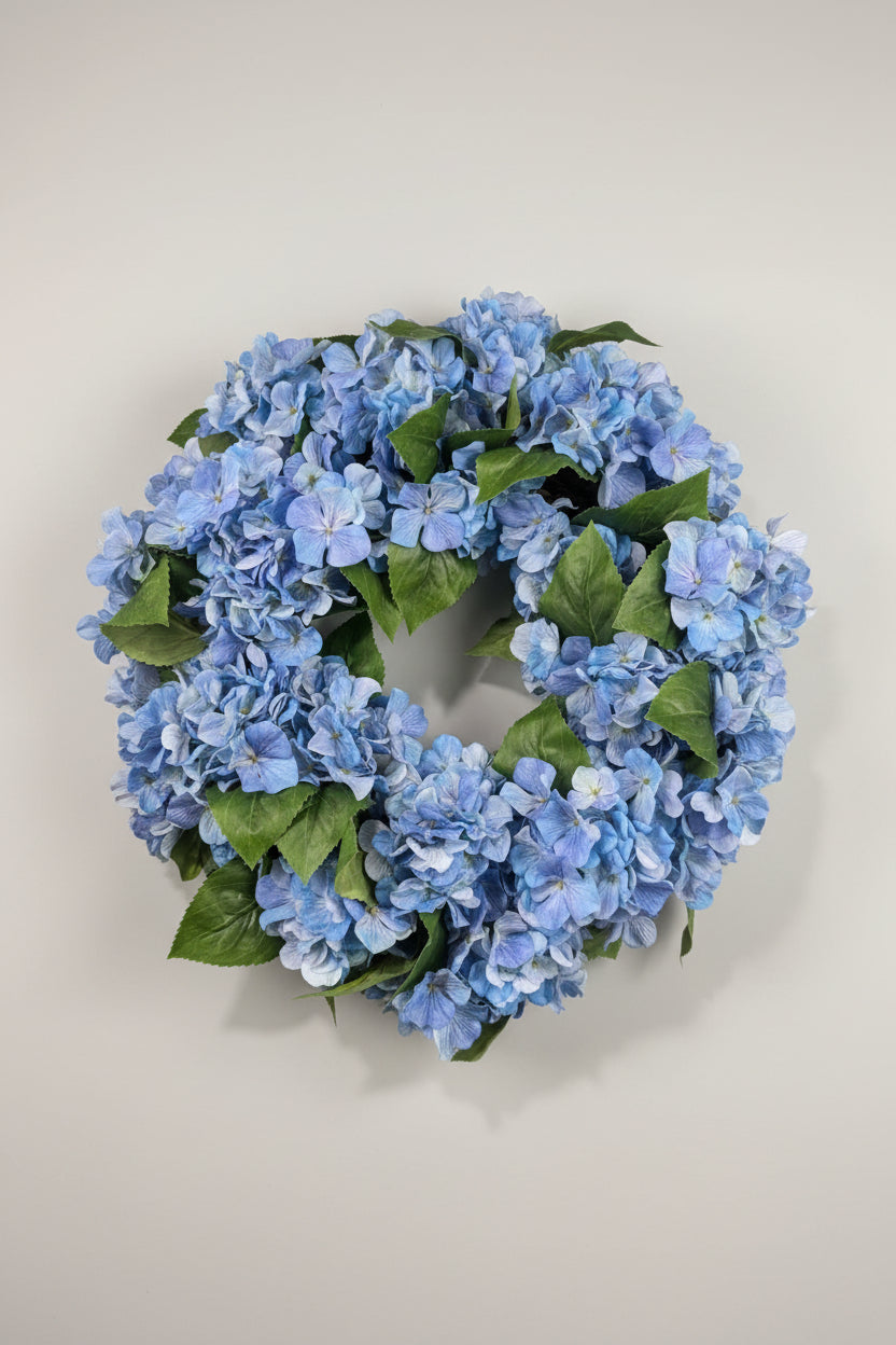 A circular artificial hydrangea wreath made of light blue flowers and green leaves, hanging on a white wall.