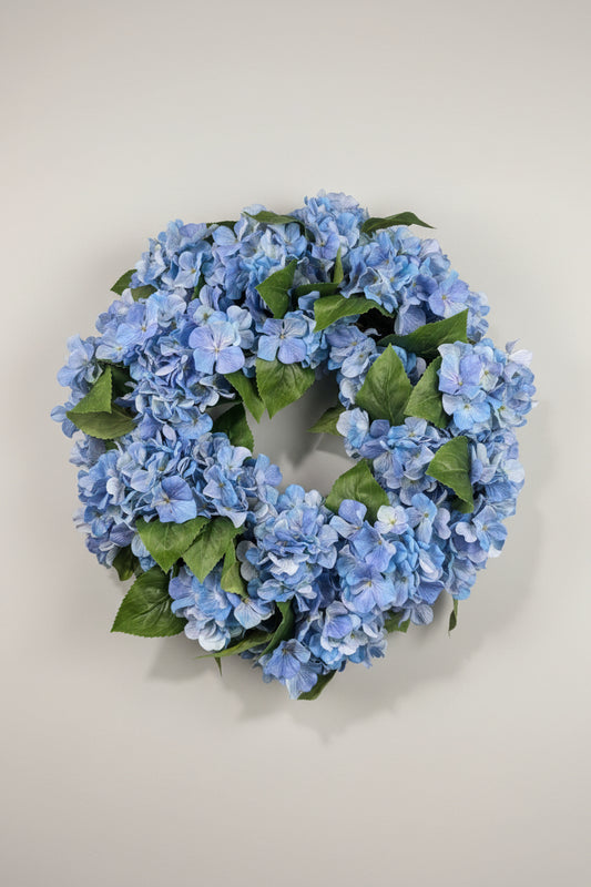A circular artificial hydrangea wreath made of light blue flowers and green leaves, hanging on a white wall.